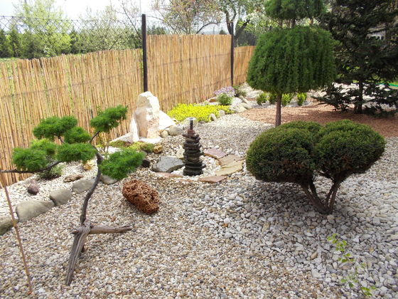 Ogród japoński.Na przodzie zdjęcia wystaje gałązka miłorzębu japońskiego,podłoże wysypane różnokolorowymi kamykami,kamienna fontanna własnoręcznie zrobiona jak i płot bambusowy